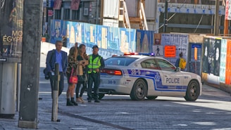 A police car is parked on a street with several people nearby, including a man in a high-visibility vest. There are construction barriers with artwork and posters, along with a sign indicating a detour for pedestrians.