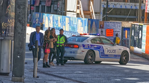 A police car is parked on a street with several people nearby, including a man in a high-visibility vest. There are construction barriers with artwork and posters, along with a sign indicating a detour for pedestrians.