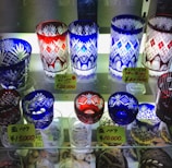A collection of intricately designed crystal glasses displayed on shelves. The glasses feature decorative patterns with floral and geometric motifs. There are various colors including red, blue, and clear glass. Price tags in Japanese yen are visible, indicating the cost of each set.