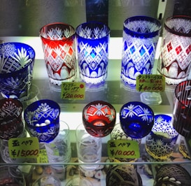 A collection of intricately designed crystal glasses displayed on shelves. The glasses feature decorative patterns with floral and geometric motifs. There are various colors including red, blue, and clear glass. Price tags in Japanese yen are visible, indicating the cost of each set.