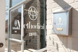 Glass door of a coffee shop with the logo and name 'Methodical Coffee' on it, alongside a metallic mailbox mounted on a brick wall.