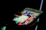 A person uses a smartphone, highlighting the WhatsApp application icon. The screen shows several other app icons with notification badges indicating unread messages.