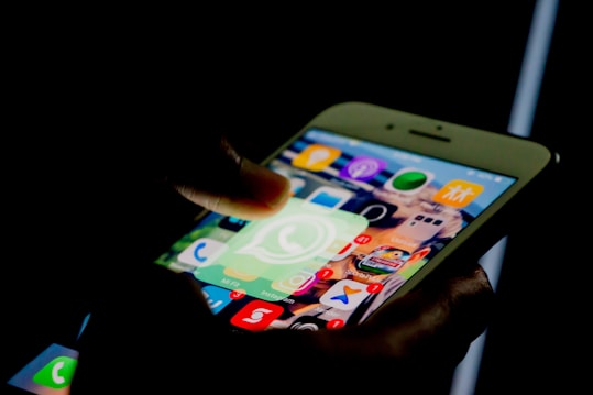 A person uses a smartphone, highlighting the WhatsApp application icon. The screen shows several other app icons with notification badges indicating unread messages.