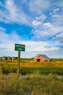 A rural landscape featuring a signpost labeled 'Snake River Canal' in the foreground, surrounded by tall grass and vegetation. In the background, a weathered red barn with a tin roof and several silo structures are visible. The sky is expansive, filled with white and gray clouds set against a rich blue backdrop.