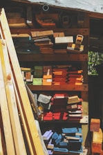A variety of wooden planks and blocks are stacked in an organized manner on shelves. The wood displays an array of colors including natural browns, vibrant oranges, blues, purples, and reds. Sunlight casts shadows, creating contrasting patterns across the wood surface.