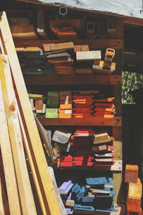 A variety of wooden planks and blocks are stacked in an organized manner on shelves. The wood displays an array of colors including natural browns, vibrant oranges, blues, purples, and reds. Sunlight casts shadows, creating contrasting patterns across the wood surface.