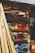 A variety of wooden planks and blocks are stacked in an organized manner on shelves. The wood displays an array of colors including natural browns, vibrant oranges, blues, purples, and reds. Sunlight casts shadows, creating contrasting patterns across the wood surface.