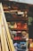 A variety of wooden planks and blocks are stacked in an organized manner on shelves. The wood displays an array of colors including natural browns, vibrant oranges, blues, purples, and reds. Sunlight casts shadows, creating contrasting patterns across the wood surface.