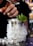 A hand is delicately adding a spoonful of sugar or salt to a metal cup filled with crushed ice and garnished with fresh mint leaves. The background is softly blurred, hinting at a bar setting.