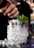 A hand is delicately adding a spoonful of sugar or salt to a metal cup filled with crushed ice and garnished with fresh mint leaves. The background is softly blurred, hinting at a bar setting.