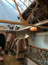 A boutique-style interior with a vintage bicycle mounted high on the wall under a glass ceiling. Below, various garments are displayed on racks, including coats and dresses. A mannequin is dressed in a colorful top. The space features decorative railings and a lit pendant lamp, creating a cozy atmosphere.