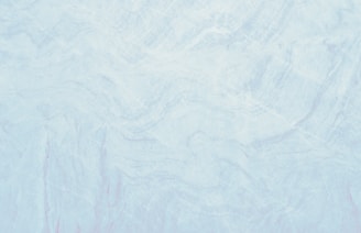 A marble texture with a smooth and subtle mix of light blue and white tones. The surface pattern consists of delicate, swirling veins and slight variations in color, resembling natural stone.