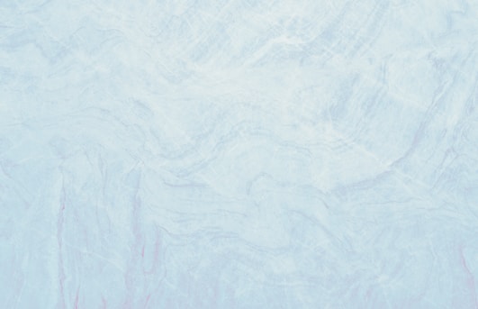 A marble texture with a smooth and subtle mix of light blue and white tones. The surface pattern consists of delicate, swirling veins and slight variations in color, resembling natural stone.