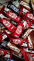 An assortment of chocolate bars with recognizable brands and packaging spread out in a dense layout. The vibrant red packages of Kit Kat bars stand out among the others, with a few Snickers and Galaxy bars in their distinct packages.