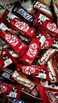 An assortment of chocolate bars with recognizable brands and packaging spread out in a dense layout. The vibrant red packages of Kit Kat bars stand out among the others, with a few Snickers and Galaxy bars in their distinct packages.