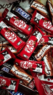 An assortment of chocolate bars with recognizable brands and packaging spread out in a dense layout. The vibrant red packages of Kit Kat bars stand out among the others, with a few Snickers and Galaxy bars in their distinct packages.