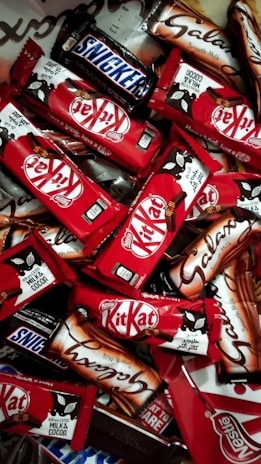 An assortment of chocolate bars with recognizable brands and packaging spread out in a dense layout. The vibrant red packages of Kit Kat bars stand out among the others, with a few Snickers and Galaxy bars in their distinct packages.