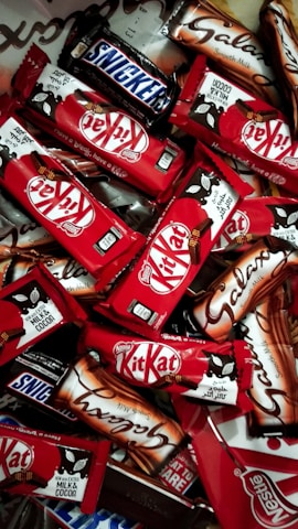 An assortment of chocolate bars with recognizable brands and packaging spread out in a dense layout. The vibrant red packages of Kit Kat bars stand out among the others, with a few Snickers and Galaxy bars in their distinct packages.