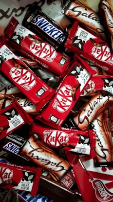 An assortment of chocolate bars with recognizable brands and packaging spread out in a dense layout. The vibrant red packages of Kit Kat bars stand out among the others, with a few Snickers and Galaxy bars in their distinct packages.