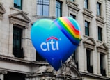 A large blue heart-shaped balloon is floating against the backdrop of a classic white stone building with multiple windows. The balloon displays the word 'Citi' in white lettering, along with a vibrant rainbow stripe along the side.