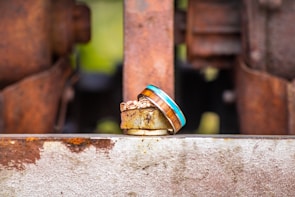A pair of rings is placed on a rusted metal surface with an industrial backdrop. One ring features a natural wood inlay bordered by a bright blue line, while the other has an intricate gold pattern.