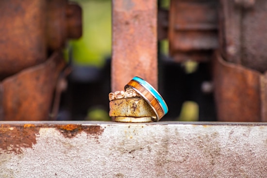 A pair of rings is placed on a rusted metal surface with an industrial backdrop. One ring features a natural wood inlay bordered by a bright blue line, while the other has an intricate gold pattern.