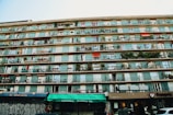 A multi-story residential building with numerous balconies, each decorated with plants and furniture. The bottom level houses various storefronts including a library and a food establishment. Some parts of the building show graffiti, adding an urban feel.