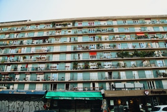 A multi-story residential building with numerous balconies, each decorated with plants and furniture. The bottom level houses various storefronts including a library and a food establishment. Some parts of the building show graffiti, adding an urban feel.