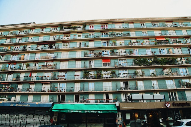 A multi-story residential building with numerous balconies, each decorated with plants and furniture. The bottom level houses various storefronts including a library and a food establishment. Some parts of the building show graffiti, adding an urban feel.