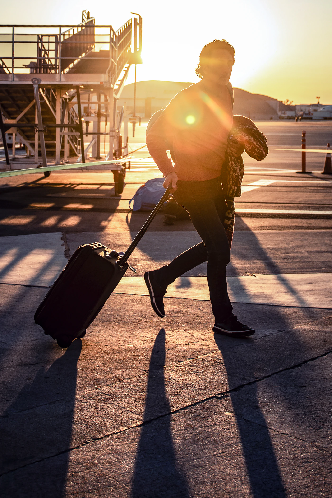 A traveler hurrying across an airport tarmac at sunset pulling a rolling suitcase near boarding stairs