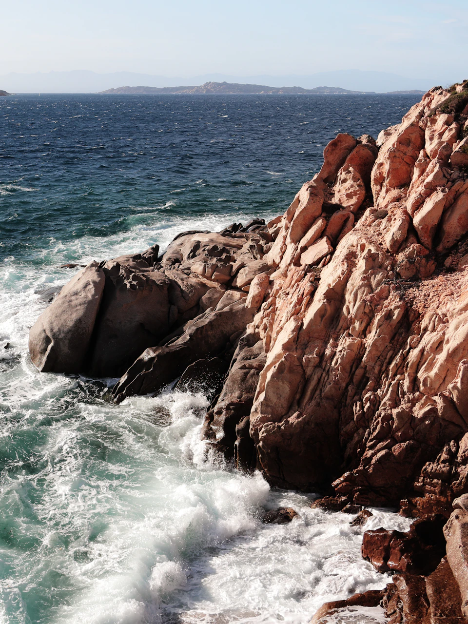 The image depicts a rocky coastline with a large, jagged rock formation on the left side and a smaller, smoother rock formation on the right side. The rocks are covered in patches of green vegetation, adding a touch of color to the otherwise stark landscape. The water surrounding the rocks is choppy, suggesting that there might be strong winds or rough seas nearby. The sky above is clear blue with a few clouds scattered across it.