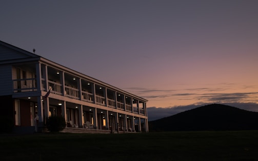 A two-story building with a row of evenly spaced lights appears at dusk. The structure features multiple balconies and appears to have a colonial architectural style. A flagpole with a flag is visible near the entrance. In the background, a hill or small mountain is silhouetted against a gradient sky transitioning from orange to purple hues.