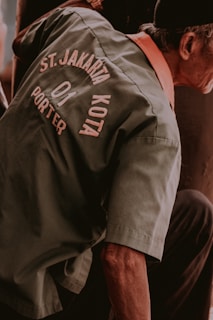 A person is wearing a green jacket with text that reads 'ST. JAKARTA KOTA PORTER' on the back. The jacket has an orange inner collar, and the person appears to be bending or sitting down.