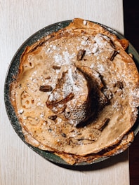 A stack of golden-brown crepes sprinkled with powdered sugar sits on a green plate. Chunks of chocolate are scattered over the top, adding a rich element to the presentation. The crepes have a light, lacy texture and are slightly curled at the edges.