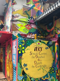 Vibrant, colorful street art covers a building wall, featuring abstract shapes, floral motifs, and patterns. A large circular design contains the text 'Art Should Comfort the Disturbed & Disturb the Comfortable' by Cesar A. Cruz. The surrounding designs include bright blues, greens, reds, and yellows with intricate details and whimsical images.