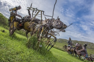 A large sculpture made from metal and mechanical parts resembles a fantastical animal, with a head similar to that of a rabbit. The sculpture is set in a grassy field under a partly cloudy sky. Another sculpture is visible in the background, also composed of metal components, depicting a human figure on a horse.