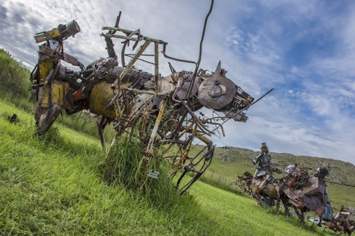 A large sculpture made from metal and mechanical parts resembles a fantastical animal, with a head similar to that of a rabbit. The sculpture is set in a grassy field under a partly cloudy sky. Another sculpture is visible in the background, also composed of metal components, depicting a human figure on a horse.