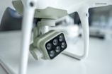 A close-up view of a camera attached to a drone, featuring multiple lenses arranged in a rectangular formation. The setting is likely a laboratory or studio, characterized by minimalist surroundings and a soft light ambience that accentuates the drone's technical components.