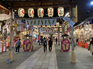 A vibrant festival setting with numerous paper lanterns overhead, stalls filled with colorful decorations, and people walking and browsing. The atmosphere is lively with a variety of items on display, creating a bustling and cultural ambiance.