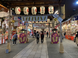 A vibrant festival setting with numerous paper lanterns overhead, stalls filled with colorful decorations, and people walking and browsing. The atmosphere is lively with a variety of items on display, creating a bustling and cultural ambiance.