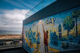 A colorful mural depicting two individuals playing basketball, with a backdrop of trees, buildings, and abstract patterns, along with the text 'THE BAY'. The scene is set against a coastline with a cloudy sky and parked cars along a road.