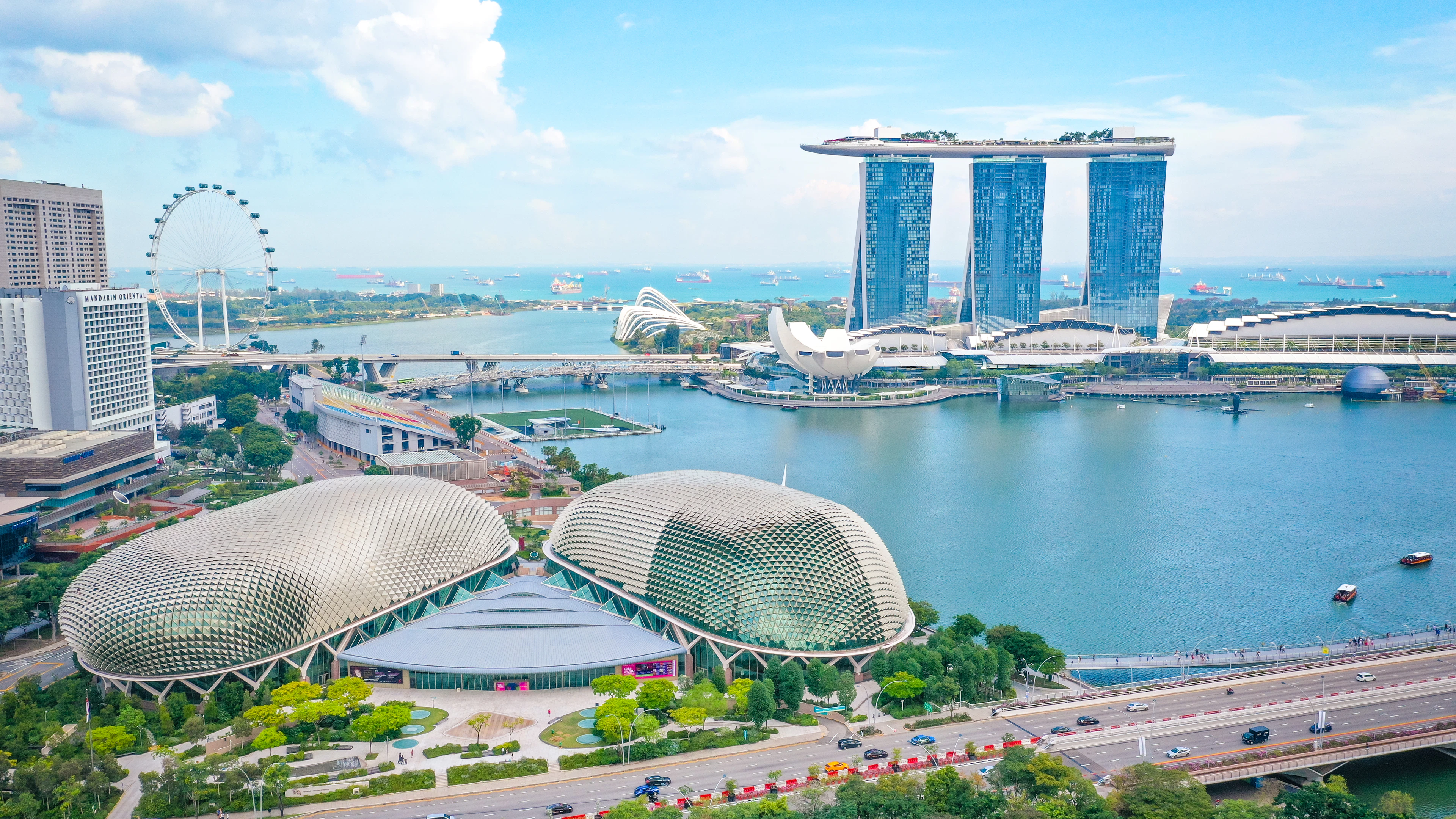 Aerial view over Marina Bay with the Singapore Flyer, Esplanade domes and Marina Bay Sands under a bright blue sky.