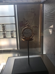 An antique pocket watch is displayed inside a glass case. The watch is open, revealing intricate dials and features. It sits on a stand, and the case is softly illuminated by overhead lighting. The background includes informational text and diagrams, adding to the historical and educational setting.