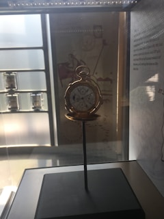 An antique pocket watch is displayed inside a glass case. The watch is open, revealing intricate dials and features. It sits on a stand, and the case is softly illuminated by overhead lighting. The background includes informational text and diagrams, adding to the historical and educational setting.