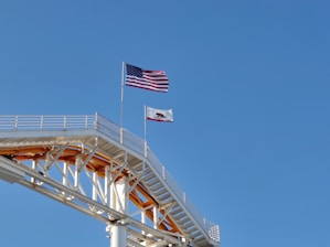 A section of a roller coaster track is visible in the foreground, with two flags waving prominently above it. The first is an American flag, and the second is the flag of California. The structure of the coaster is painted white with some yellow accents, and it contrasts against the clear blue sky.