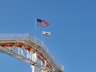 A section of a roller coaster track is visible in the foreground, with two flags waving prominently above it. The first is an American flag, and the second is the flag of California. The structure of the coaster is painted white with some yellow accents, and it contrasts against the clear blue sky.