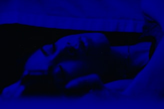 A person is lying down in a dimly lit environment with a deep blue tint. Their eyes are closed, suggesting they might be sleeping or resting. The lighting creates a soft, serene atmosphere.