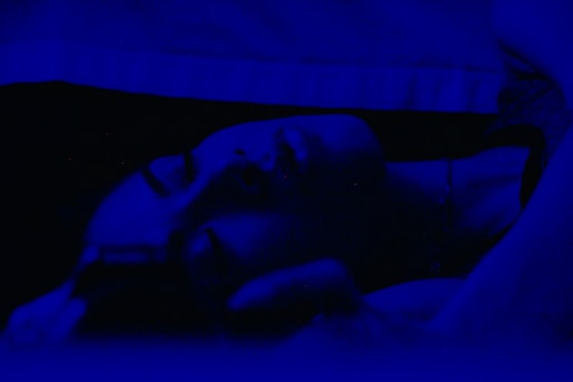A person is lying down in a dimly lit environment with a deep blue tint. Their eyes are closed, suggesting they might be sleeping or resting. The lighting creates a soft, serene atmosphere.