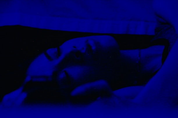 A person is lying down in a dimly lit environment with a deep blue tint. Their eyes are closed, suggesting they might be sleeping or resting. The lighting creates a soft, serene atmosphere.