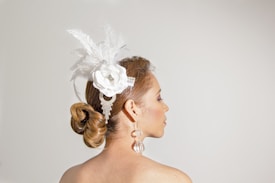 A woman with her back turned, featuring a hairstyle composed of an intricate bun adorned with white feathers and a large floral embellishment. She wears elaborate crystal earrings and her hair is styled neatly.